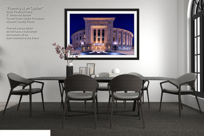 a modern dining room with a large framed picture of a stadium on the wall. The picture is framed in black and has a minimalist design. The dining table is surrounded by four chairs, and there is a vase of flowers on the table. The overall setting appears to be a sophisticated and elegant space.