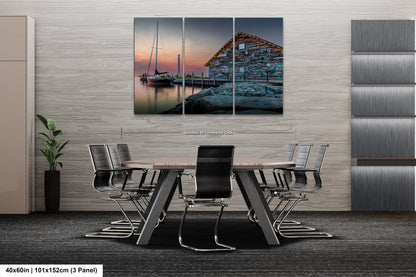 The image is a modern office space with a large, three-panel artwork depicting a serene scene of a boat on a body of water at sunset. The artwork is mounted on a gray wall above a wooden table surrounded by chairs. The overall design of the space is sleek and contemporary, with a neutral color palette and minimalist furniture.
