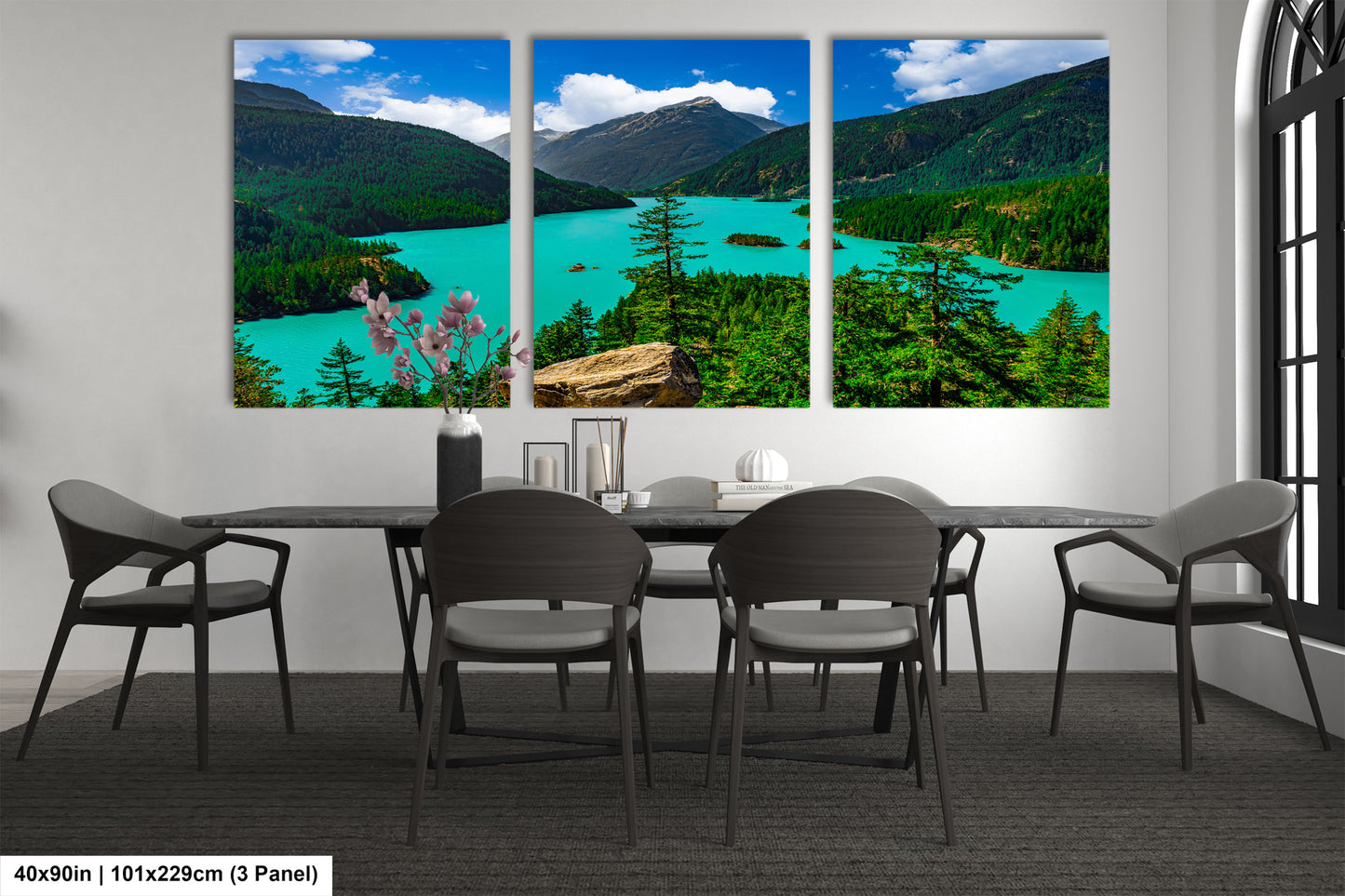 The image is a large, three-panel landscape painting depicting a scenic mountain lake surrounded by lush forests. The painting is hung on a wall above a dining table with chairs.