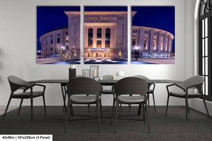 A triptych of a large, illuminated image of Yankee Stadium at night, with the stadium's name prominently displayed. The stadium is surrounded by a large, circular driveway, and the night sky is visible above. The image is framed by a white wall, and there is a dining table with chairs in front of it.
