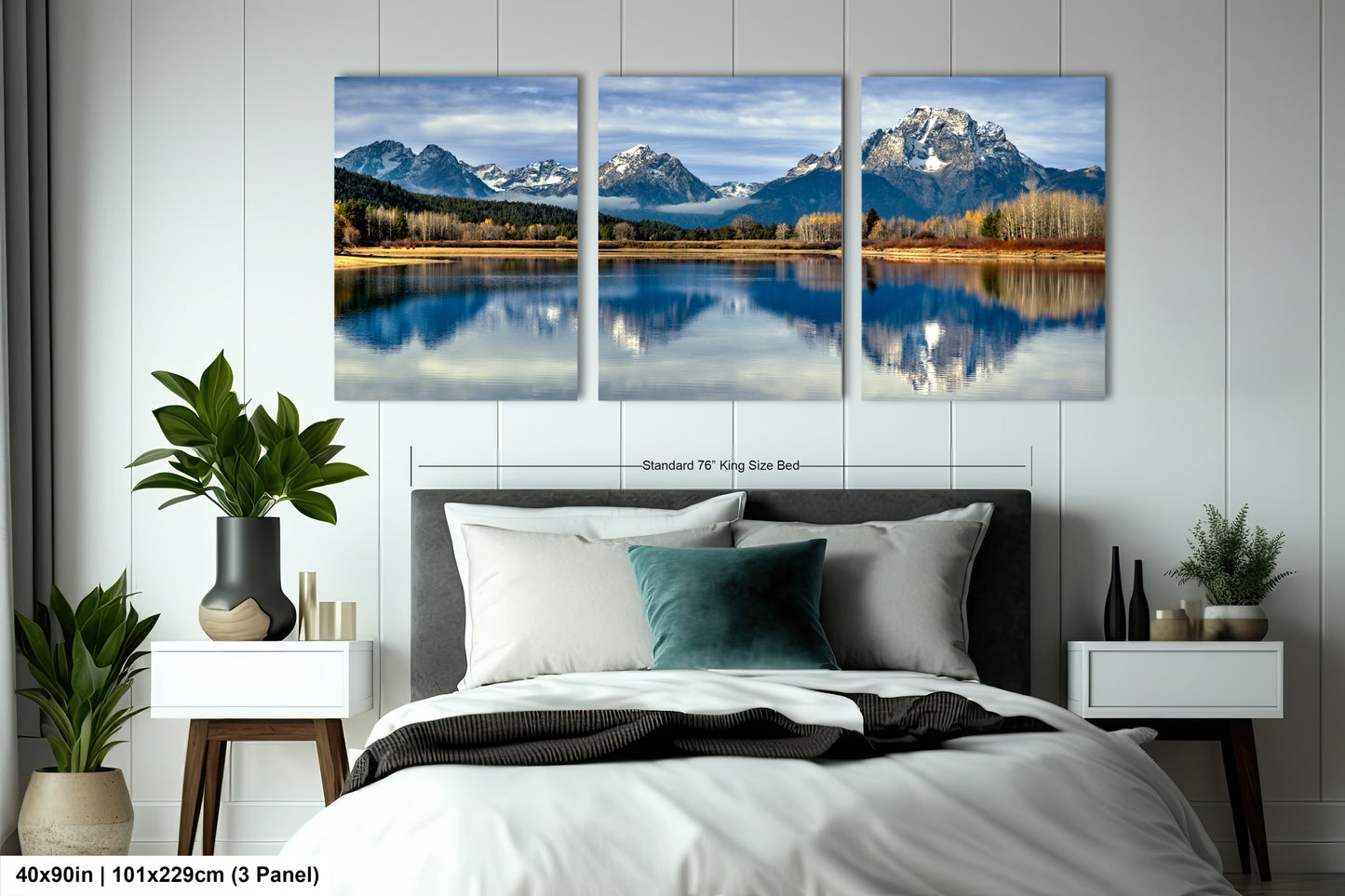 The image is a triptych of a serene landscape featuring a mountain lake, with the mountains reflected in the calm waters. The triptych is framed and displayed on a wall above a bed with white bedding and a gray headboard.