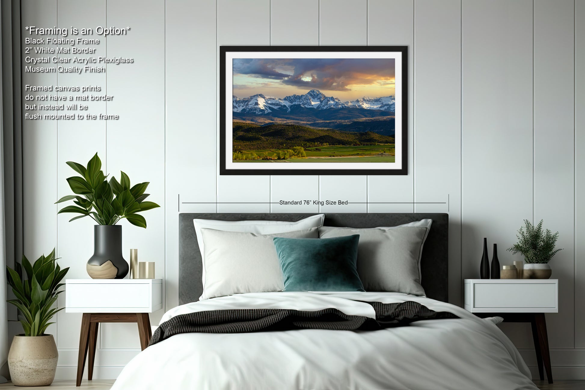 a bedroom with a large framed landscape painting hanging on the wall above a bed. The painting depicts a scenic mountain range with a cloudy sky. The room has a minimalist design with white walls, a gray headboard, and a black and white color scheme. There are also two nightstands with plants and decorative items, and a vase on one of the nightstands.