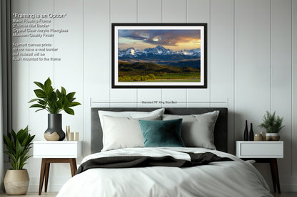 a bedroom with a large framed landscape painting hanging on the wall above a bed. The painting depicts a scenic mountain range with a cloudy sky. The room has a minimalist design with white walls, a gray headboard, and a black and white color scheme. There are also two nightstands with plants and decorative items, and a vase on one of the nightstands.
