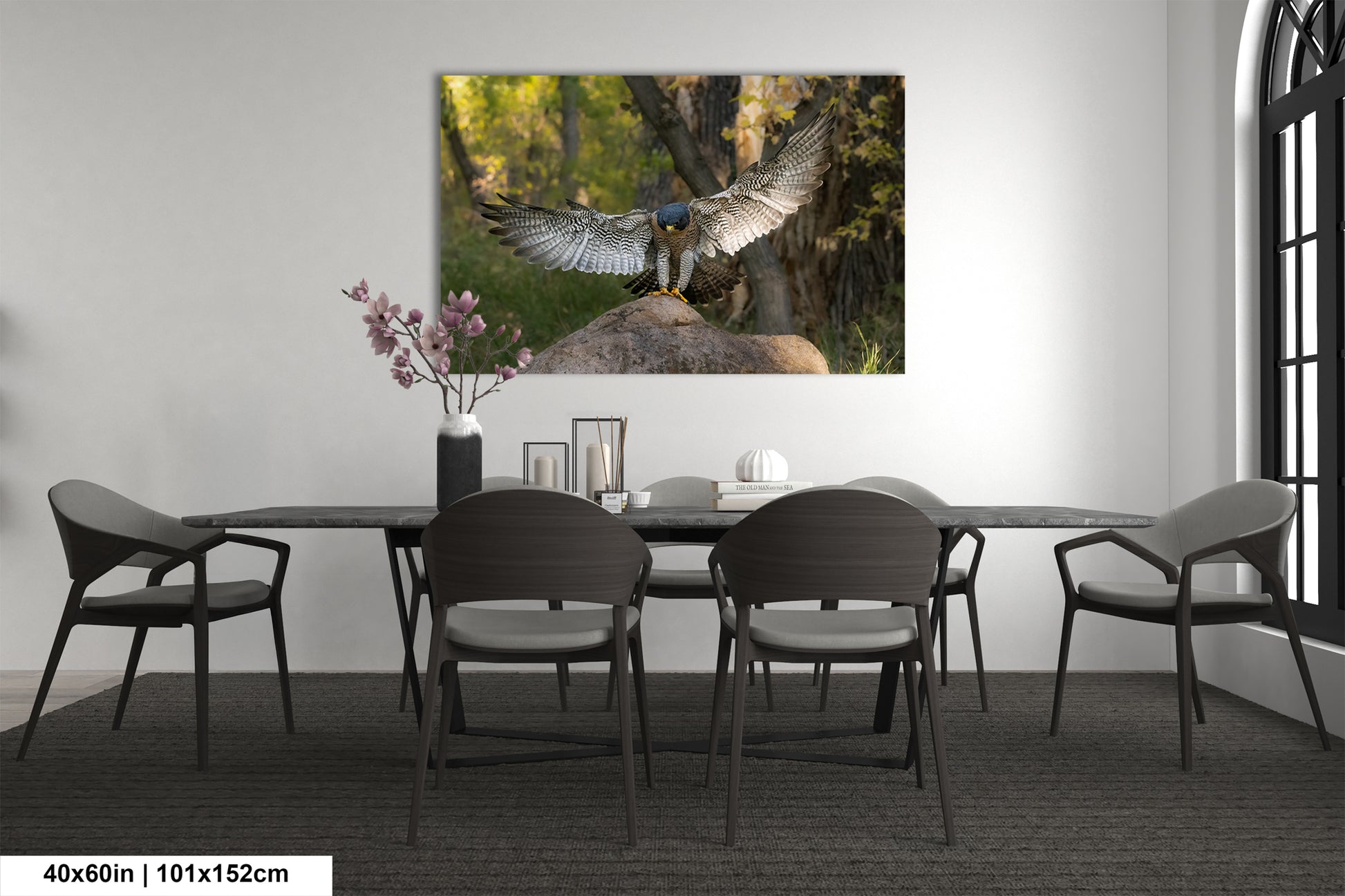 a large bird of prey, possibly a hawk or falcon, with its wings spread wide, perched on a rock in a forest setting. The bird is the focal point of the image, which is displayed on the wall above a dining table with four chairs.