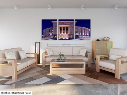 The image is a modern living room with a large, illuminated stadium in the background. The room is furnished with a sofa, two chairs, and a coffee table. The furniture appears to be made of wood and has a neutral color scheme. The room has a minimalist and clean design, with a geometric patterned rug on the floor.