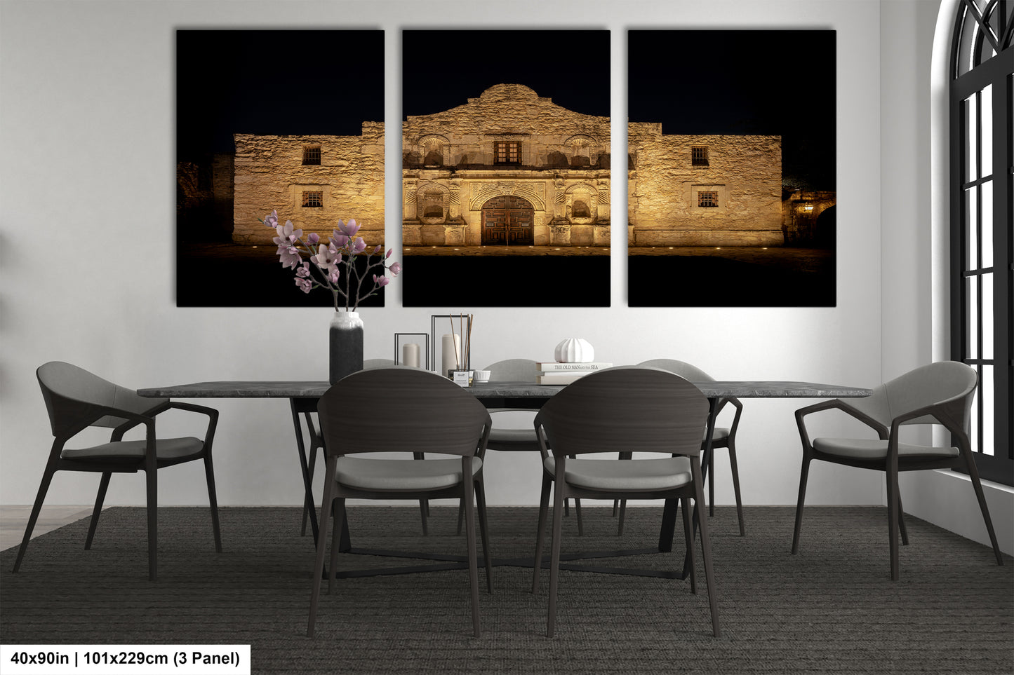 a modern dining room with a large, illuminated photograph of the historic Alamo in the background. The room is furnished with a dining table and chairs, and there is a vase of flowers on the table.