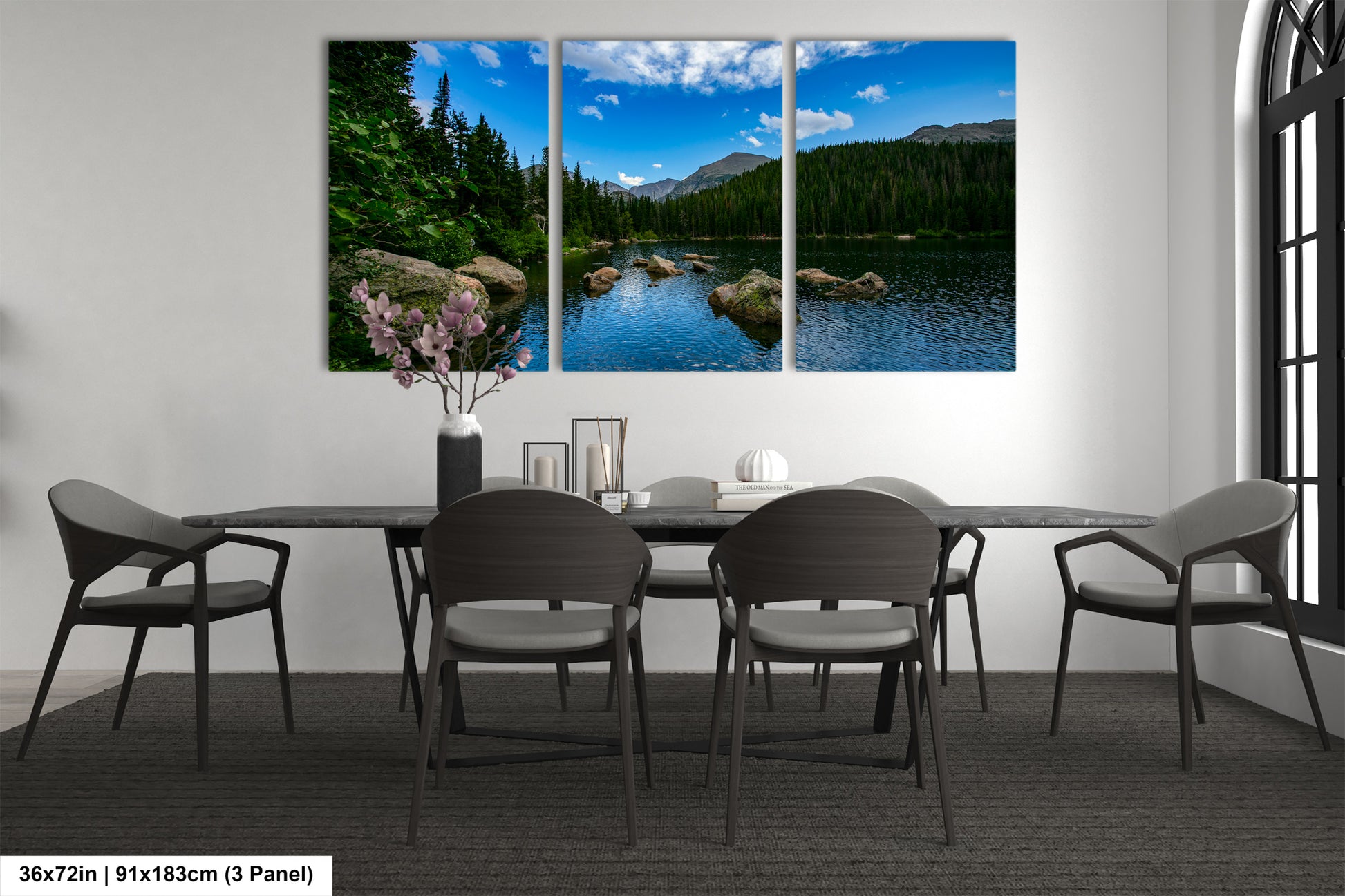 a modern dining room with a large, three-panel landscape painting depicting a serene mountain lake surrounded by trees. The painting is hung on the wall above a black dining table with six chairs. The room has a minimalist and contemporary design, with a vase of flowers and a few decorative items on the table.
