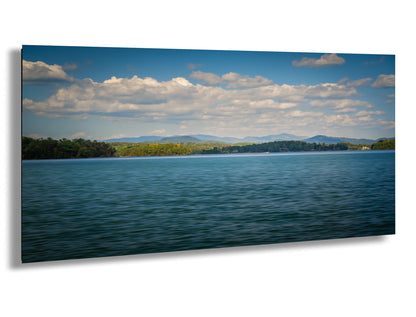 a serene lake surrounded by lush green trees and mountains under a blue sky with fluffy white clouds.