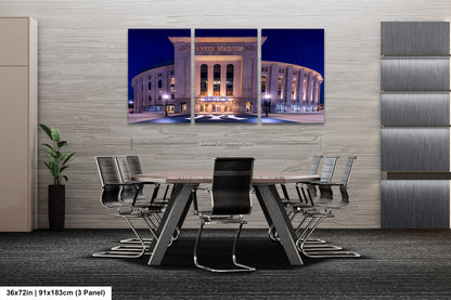 A modern office space with a large, circular building in the background, likely a stadium or arena, illuminated at night. The office has a conference table surrounded by chairs, and there is a plant in the corner.