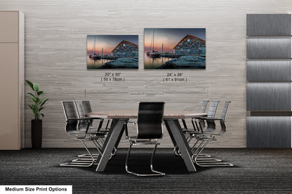 a modern office space with a large wooden table surrounded by black chairs. Above the table, there are two framed prints of a seascape, one depicting a sailboat and the other showing a building. The prints are of different sizes, with the larger one being 24" x 36" and the smaller one being 20" x 30". The prints are displayed on a wall, and there is a plant in the corner of the room.