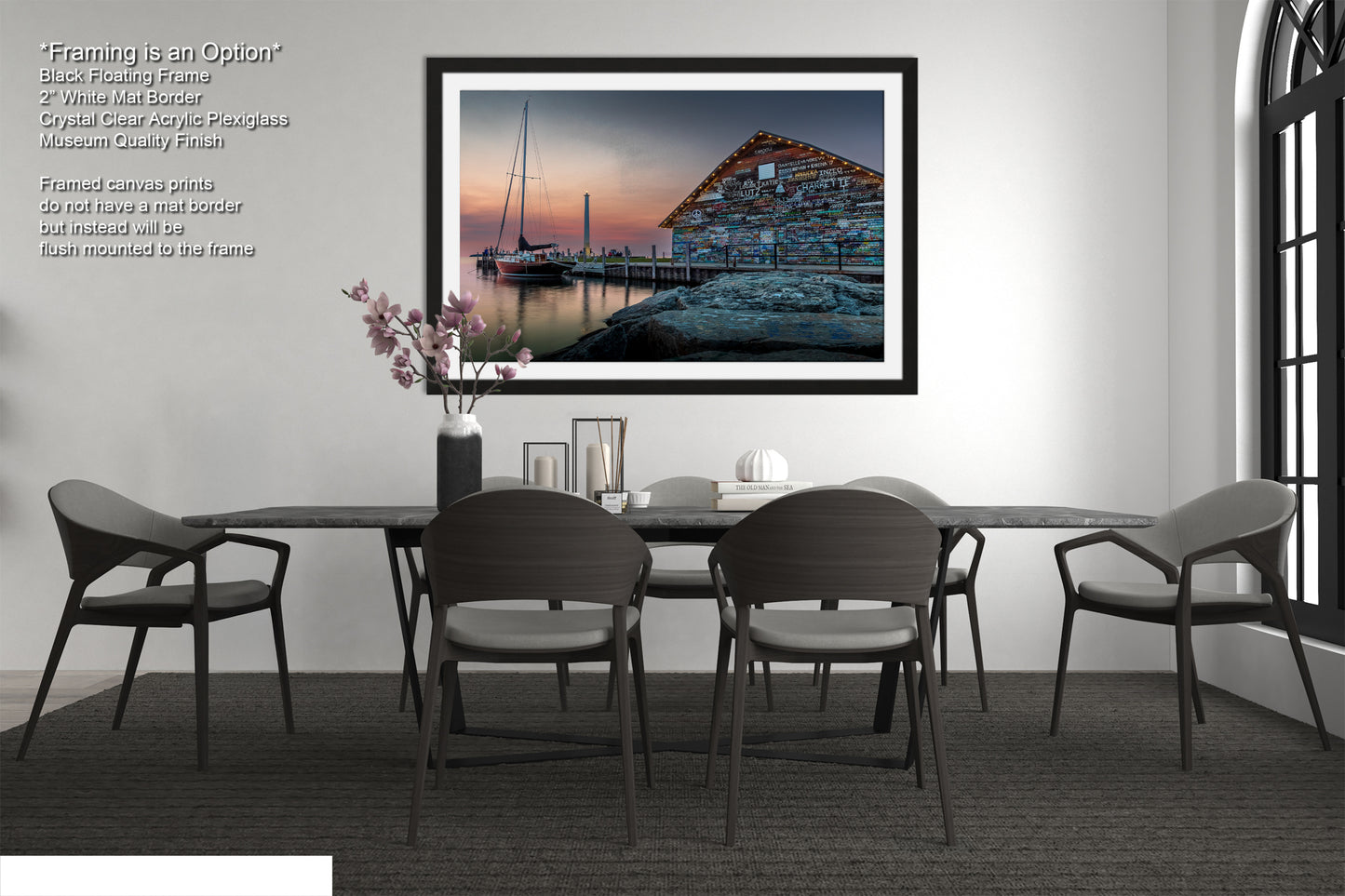 a modern dining room with a large black dining table surrounded by six matching chairs. Above the table, there is a framed photograph of a building with a dock and a sailboat in the background, creating a serene and picturesque scene. The room has a minimalist and contemporary design, with a white wall and a window providing natural light.