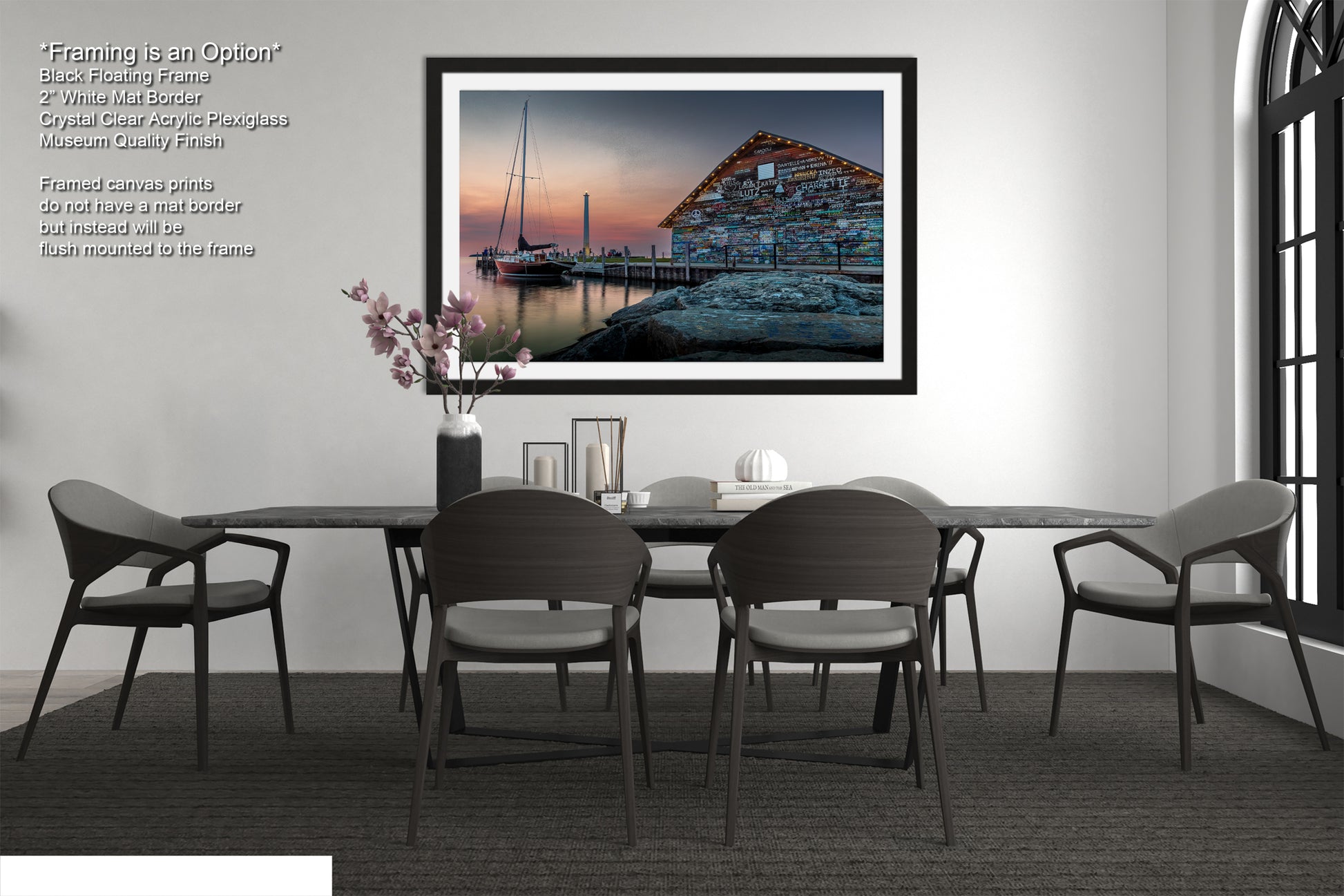 a modern dining room with a large black dining table surrounded by six matching chairs. Above the table, there is a framed photograph of a building with a dock and a sailboat in the background, creating a serene and picturesque scene. The room has a minimalist and contemporary design, with a white wall and a window providing natural light.