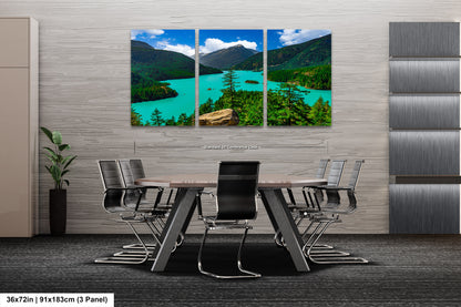 A modern office space with a large, three-panel landscape painting depicting a scenic mountain lake surrounded by forests. The painting is mounted on a gray wall above a wooden table with chairs.