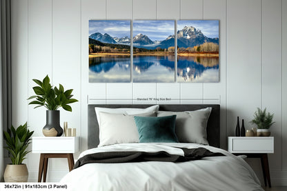 A bedroom with a large, three-panel landscape painting of a mountain lake hanging on the wall above the bed. The painting features a serene scene of a lake surrounded by mountains, with the mountains reflecting the sky and the lake's surface. The room is decorated with potted plants, a vase, and other furnishings, creating a cozy and inviting atmosphere.