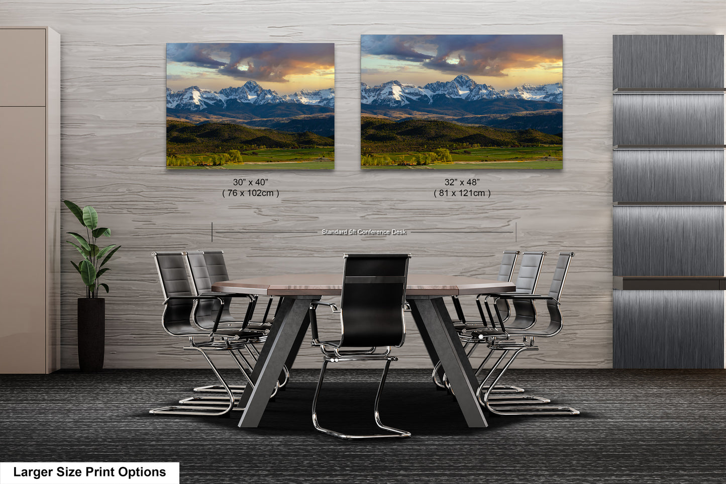 a modern office space with a large wooden table surrounded by black chairs. Two framed landscape paintings are displayed on the wall above the table, depicting a scenic mountain landscape.
