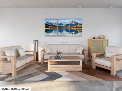 A modern living room with a large, three-panel landscape painting depicting a serene mountain lake scene. The painting is hung on the wall above a wooden coffee table, which is surrounded by two white couches and a matching chair. The room has a minimalist and clean design, with a neutral color palette and a geometric patterned rug on the floor.