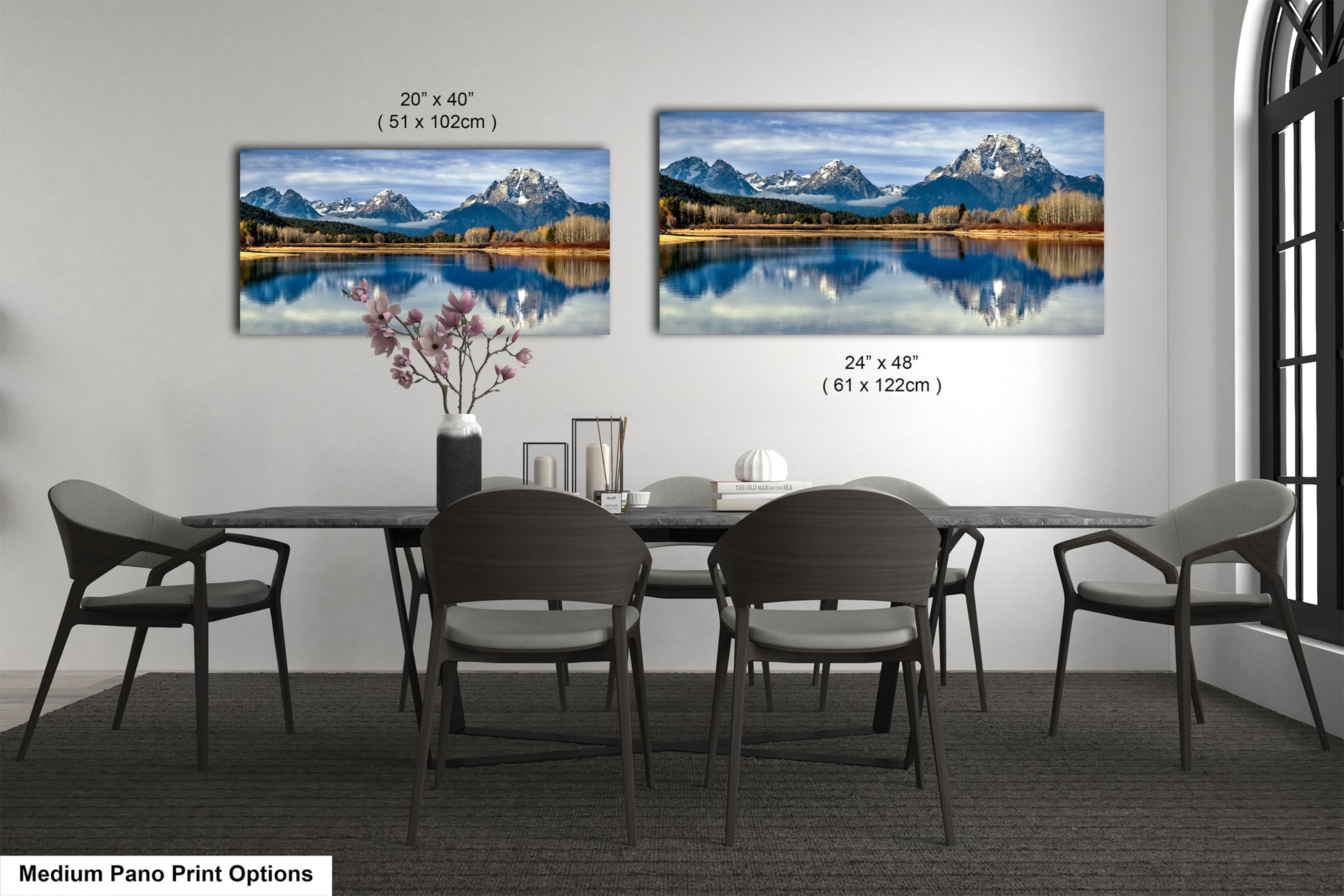 a modern dining room with a large pano print of a scenic mountain landscape on the wall, featuring a lake and snow-capped peaks. The print is framed in a medium size, with dimensions of 24" x 48" (51 x 102 cm) and 24" x 48" (51 x 102 cm). The dining table is surrounded by chairs, and there is a vase of flowers and a book on the table.