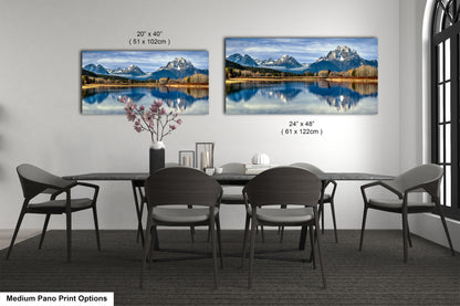 a modern dining room with a large pano print of a scenic mountain landscape on the wall, featuring a lake and snow-capped peaks. The print is framed in a medium size, with dimensions of 24" x 48" (51 x 102 cm) and 24" x 48" (51 x 102 cm). The dining table is surrounded by chairs, and there is a vase of flowers and a book on the table.