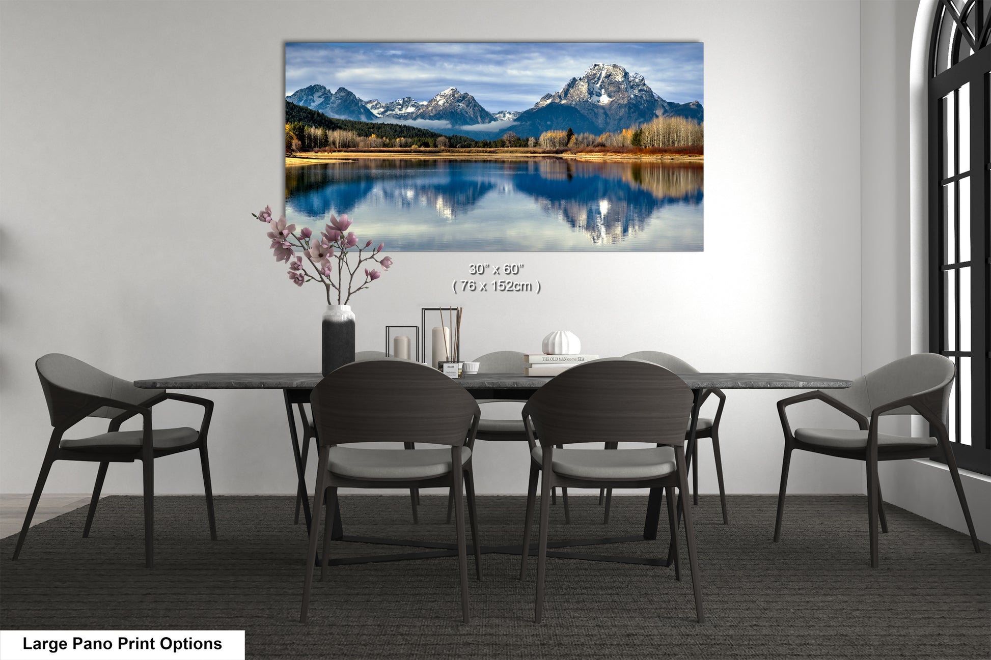 A large pano print depicting a serene mountain landscape with a lake and snow-capped peaks, hanging on a wall above a dining table with chairs.