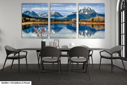a modern dining room with a large panoramic view of a mountain lake and snow-capped mountains in the background. The room is furnished with a dining table and chairs, and there is a vase of flowers on the table. The overall design and decor create a serene and tranquil atmosphere.