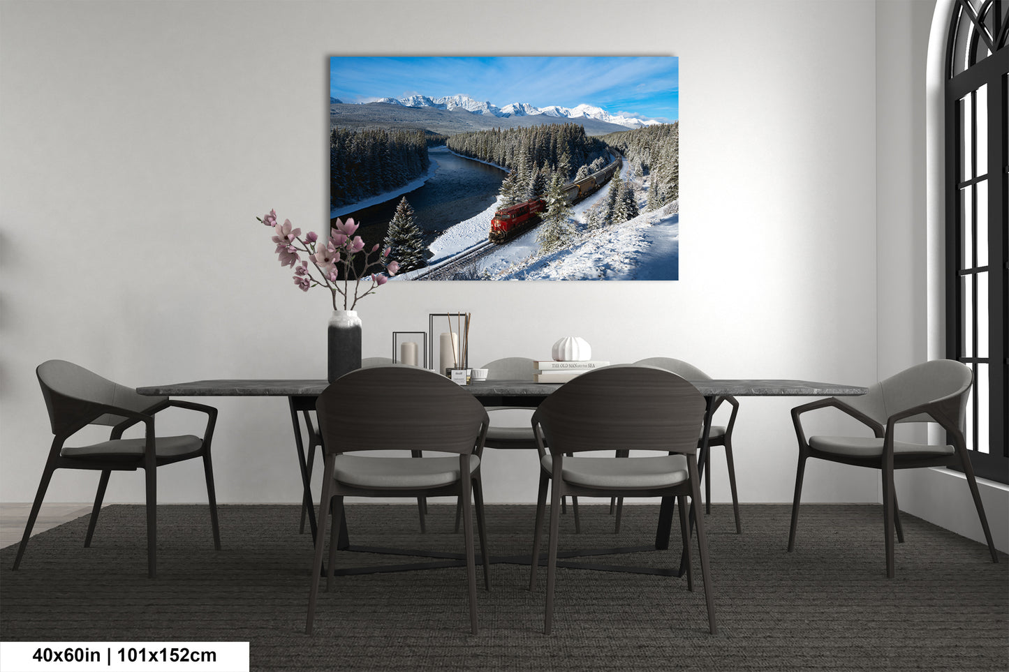 a large, framed picture of a snowy mountain landscape with a train passing through it, hanging on a wall above a dining table with chairs.