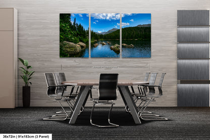 A modern office space with a large, three-panel landscape painting depicting a serene lake surrounded by mountains and trees. The painting is mounted on a gray wall above a wooden table with chairs.