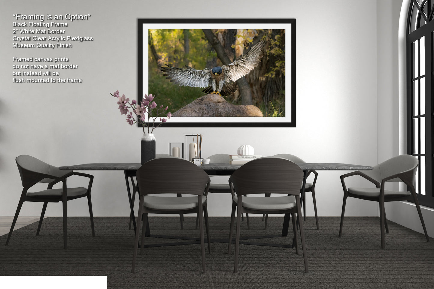 a modern dining room with a large framed photograph of a bird of prey, possibly a hawk or falcon, perched on a rock in a forest setting. The photograph is framed in black and hangs on the wall above a black dining table surrounded by matching chairs. The room has a minimalist and contemporary design, with a vase of flowers, a book, and a bottle of wine on the table, creating a serene and elegant atmosphere.