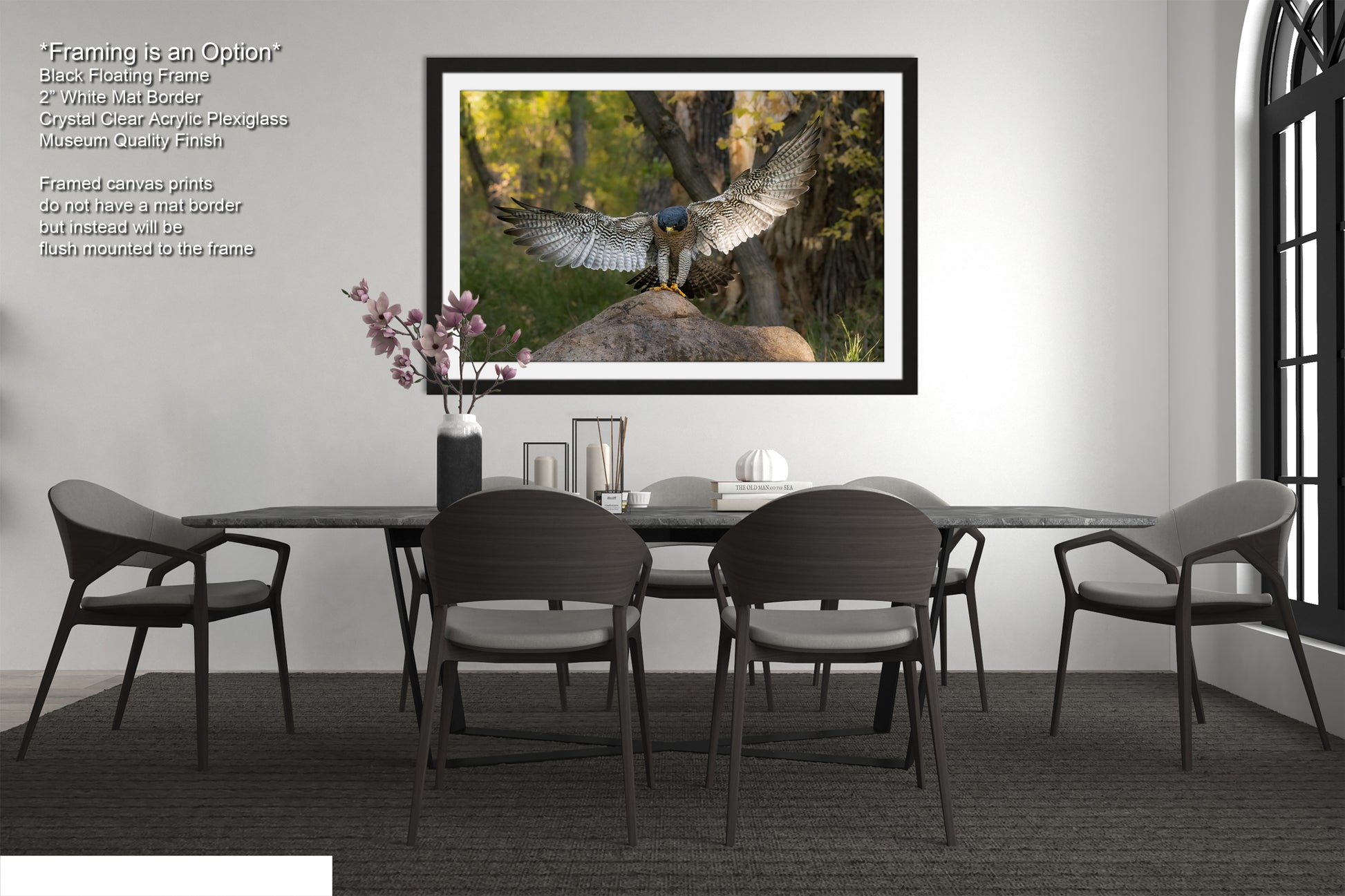 a modern dining room with a large framed photograph of a bird of prey, possibly a hawk or falcon, perched on a rock in a forest setting. The photograph is framed in black and hangs on the wall above a black dining table surrounded by matching chairs. The room has a minimalist and contemporary design, with a vase of flowers, a book, and a bottle of wine on the table, creating a serene and elegant atmosphere.