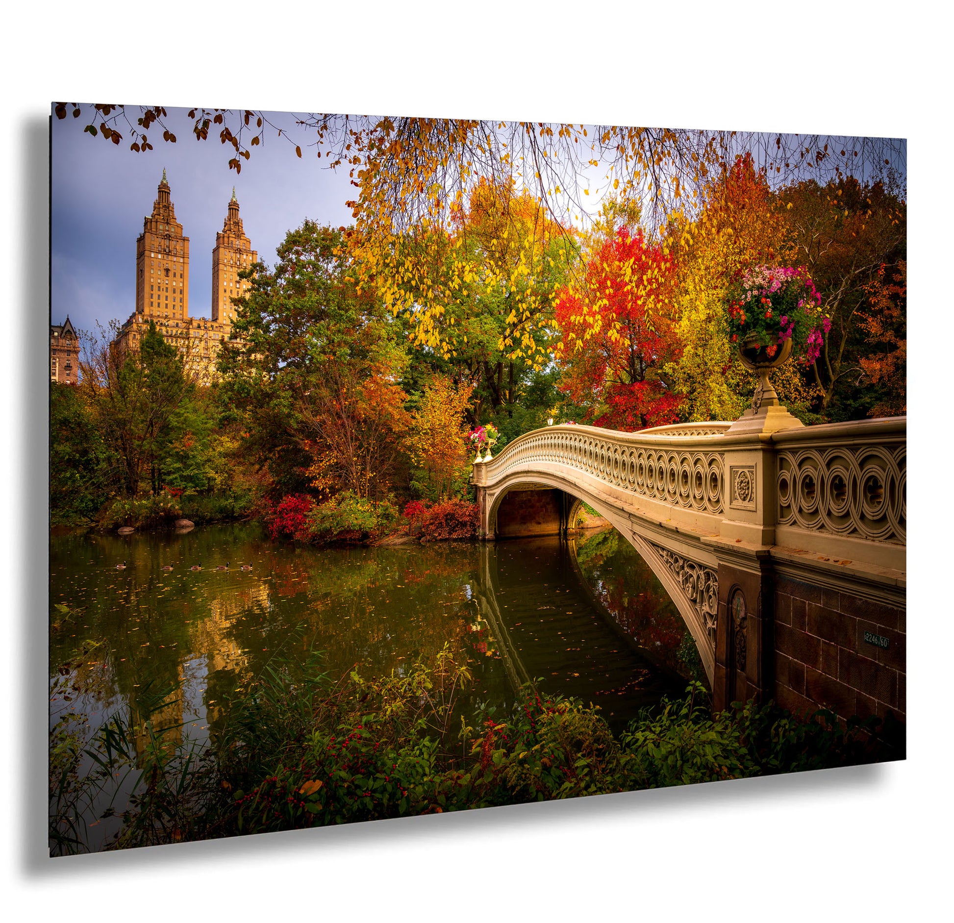 a picturesque scene of a stone bridge spanning a tranquil pond, surrounded by a vibrant display of autumn foliage. The bridge is adorned with flowers, adding a touch of elegance to the scene. In the background, the iconic New York City skyline, including the famous Central Park, can be seen, creating a beautiful contrast between the natural beauty of the park and the architectural marvel of the city.