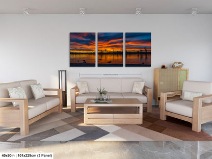 The image is a modern living room with a large, three-panel landscape painting hanging on the wall. The painting depicts a serene sunset over a body of water, with the sky filled with vibrant colors. The room is furnished with a sofa, two chairs, and a coffee table, creating a comfortable and inviting atmosphere.