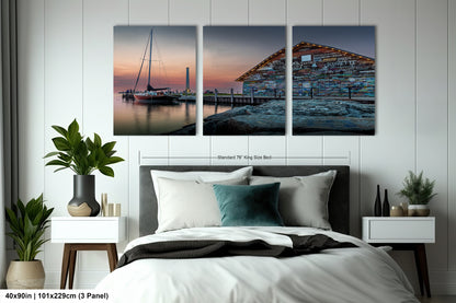 a modern office space with a large, three-panel artwork depicting a serene harbor scene at sunset. The artwork is mounted on a gray wall above a wooden table surrounded by black chairs. The overall design of the space is sleek and contemporary, with a neutral color palette and minimalist furniture.