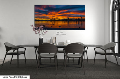 A large pano print depicting a sunset over a body of water, with silhouettes of trees and birds visible in the sky.