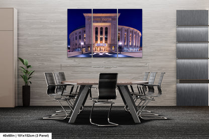 The image is a modern office space with a large, illuminated photograph of Yankee Stadium in New York City displayed on the wall. The photograph is composed of three panels, and the image is framed with a black border. The office has a minimalist design, featuring a gray wall, a wooden floor, and a black chair in front of a wooden desk.