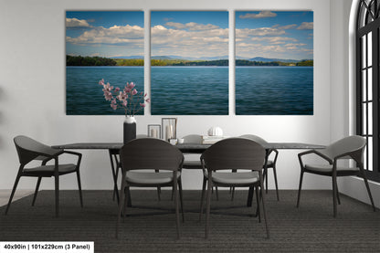 a modern dining room with a large window overlooking a serene lake. The room is furnished with a black dining table surrounded by six chairs, and there is a vase of flowers on the table. The wall behind the table displays three large panoramic paintings depicting a picturesque lake scene with mountains in the distance.