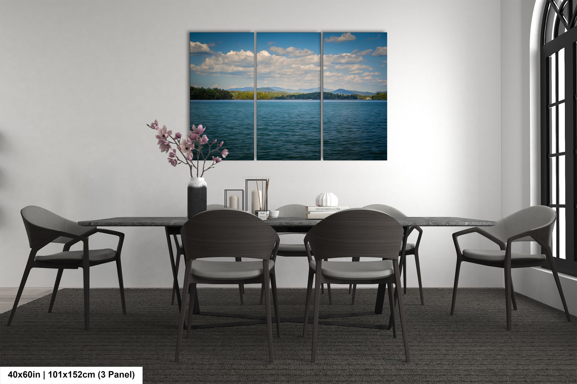 a modern dining room with a large, three-panel landscape painting of a serene lake and sky above a mountain range. The painting is hung on the wall above a black dining table surrounded by six chairs. The room has a minimalist and elegant design, with a vase of pink flowers and a book on the table adding a touch of color and personality to the space.