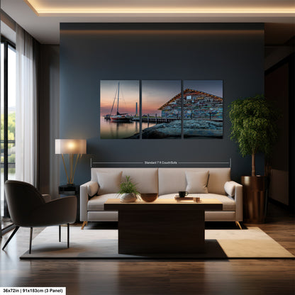 The image is a modern dining room with a large wall-mounted painting depicting a serene scene of a boat on a body of water at sunset. The painting is composed of three panels, each capturing different aspects of the serene landscape. The dining table is surrounded by four chairs, and a vase of flowers adds a touch of elegance to the room. The overall design and arrangement of the furniture and artwork create a harmonious and inviting atmosphere.