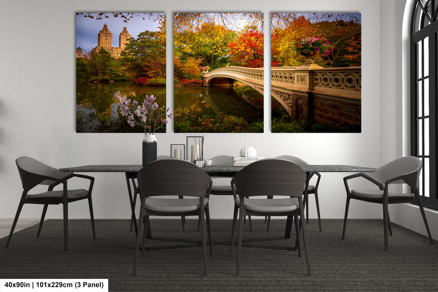 The image is a modern dining room with a large wall-mounted painting depicting a scenic view of Central Park in New York City. The painting features a bridge, trees, and a lake, creating a serene and picturesque atmosphere. The dining table is surrounded by chairs, and there are decorative elements such as a vase and a book on the table. The overall design and layout of the room suggest a stylish and comfortable dining experience.