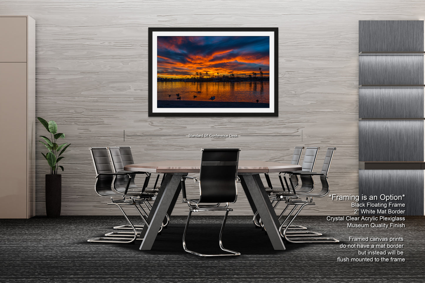a modern office space with a large framed artwork depicting a sunset over a body of water. The artwork is framed in a black floating frame and is mounted on a gray wall above a wooden table surrounded by black chairs. The office has a minimalist design with gray walls and a gray carpeted floor.