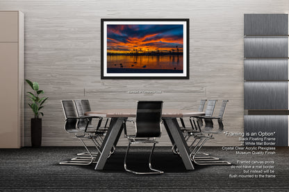 a modern office space with a large framed artwork depicting a sunset over a body of water. The artwork is framed in a black floating frame and is mounted on a gray wall above a wooden table surrounded by black chairs. The office has a minimalist design with gray walls and a gray carpeted floor.