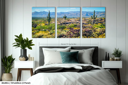 The image is a bedroom with a large bed, nightstands, and potted plants. Above the bed, there is a large canvas print depicting a desert landscape with cacti and mountains in the background.