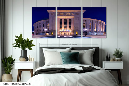 The image is a triptych of a large stadium at night, featuring the exterior of Yankee Stadium. The stadium is illuminated, showcasing its distinctive architecture. The triptych is framed and displayed on a wall in a bedroom setting, with a bed and nightstand visible in the foreground.