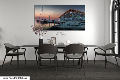 A large pano print depicting a serene harbor scene at sunset, with a wooden building and a sailboat reflected in the calm waters.
