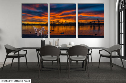 a modern dining room with a large, three-panel wall art depicting a serene sunset over a body of water. The art piece is framed in a black frame and hangs on a white wall above a black dining table surrounded by gray chairs. The table is set with a vase of flowers and a few decorative items, creating a cozy and inviting atmosphere.