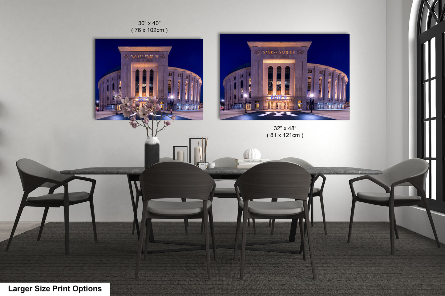 a large-sized print of a building at night, with two smaller prints of the same building displayed on the wall above it. The print on the wall is larger than the one on the table, and both prints are framed. The table is surrounded by chairs, creating a dining area.