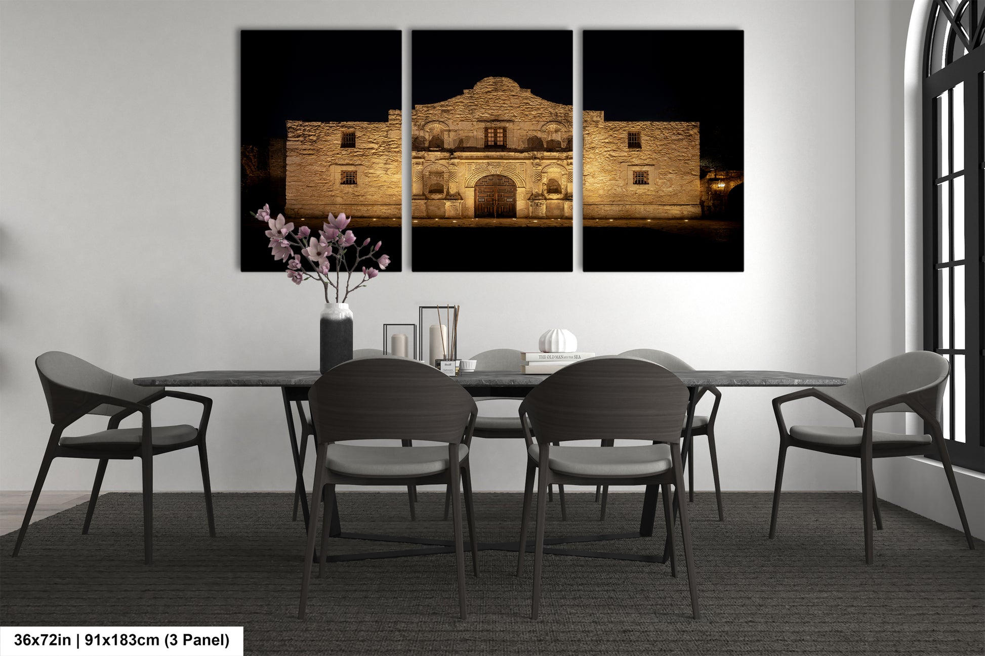 a modern dining room with a large, illuminated painting of the Alamo in the background. The painting is divided into three panels, creating a sense of depth and perspective. The room is furnished with a black table and chairs, and a vase of flowers adds a touch of color to the otherwise monochrome setting.