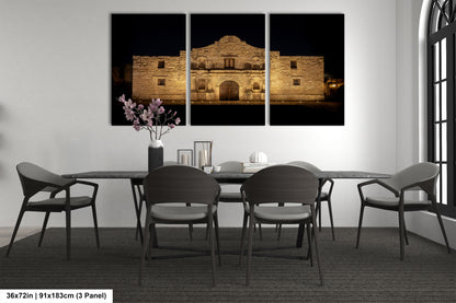 a modern dining room with a large, illuminated painting of the Alamo in the background. The painting is divided into three panels, creating a sense of depth and perspective. The room is furnished with a black table and chairs, and a vase of flowers adds a touch of color to the otherwise monochrome setting.