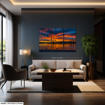 a modern living room with a large, three-panel landscape painting hanging on the wall. The painting features a sunset over a body of water, with silhouettes of trees and buildings visible in the background. The room is furnished with a white sofa, a coffee table, and a potted plant, creating a cozy and inviting atmosphere.