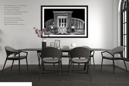 a modern dining room with a large framed black and white photograph of a stadium on the wall. The photograph is framed in a black frame and is the focal point of the room. The dining table is surrounded by four chairs, and there is a vase of flowers on the table. The room has a minimalist and clean design, with white walls and a gray carpet.