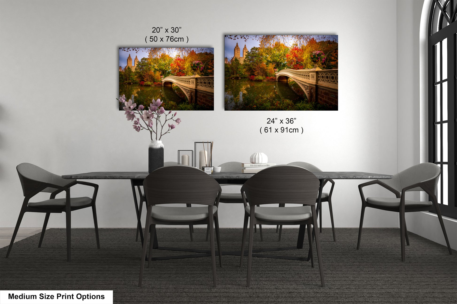 a dining room with a long table and six chairs. There are two framed prints hanging on the wall above the table, depicting a scenic view of a bridge and trees. The prints are of different sizes, with one being larger than the other. The table is set with a vase of flowers and a book, creating a cozy and inviting atmosphere.