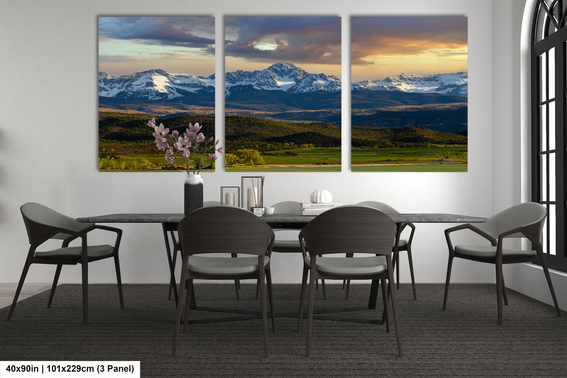 a dining room with a large panoramic view of a mountain range in the background. The room has a black table and chairs, and a vase of flowers on the table. The wall behind the table displays three large paintings depicting the mountain range and the sky.